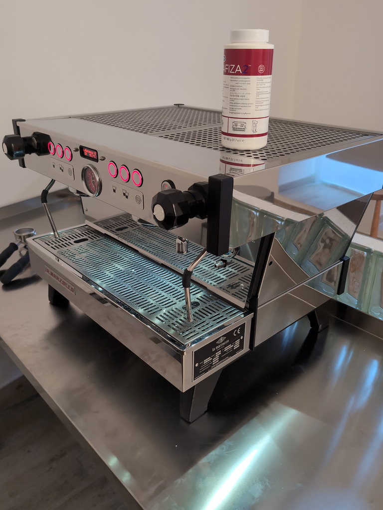 Fotografie zobrazuje profesionální pákový kávovar značky La Marzocco, model Linea Mini, známý svou spolehlivostí a vysokou kvalitou v kavárenském prostředí. Kávovar je vyfocen na nerezovém pultu, na vrchní části je nádoba čisticího prostředku Cafiza 2, určeného k údržbě a čištění skupinových hlav.
