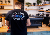 Záda hlavního technika v černém firemním potištěném tričku nad kávovarem Synesso v kavárně.